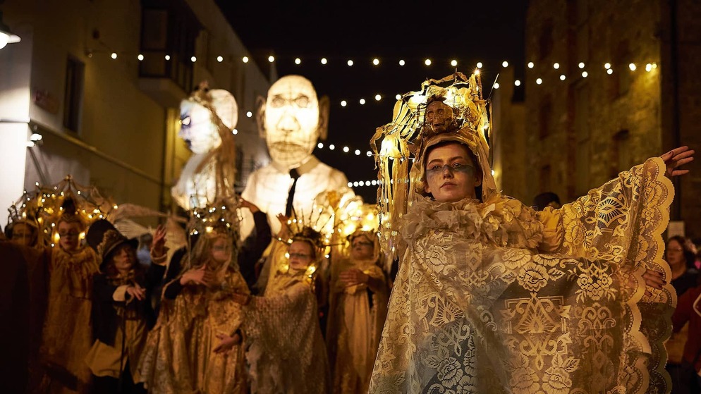Performer in ornate lace costume with glowing headpiece leads night-time Halloween parade with giant lantern puppets in Limerick.