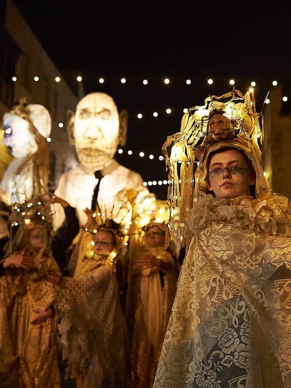 Performer in ornate lace costume with glowing headpiece leads night-time Halloween parade with giant lantern puppets in Limerick.