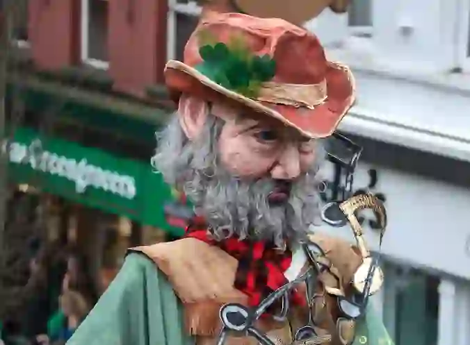 Costumed street performer with a large mask, shamrock hat and musical notes at a St Patrick’s Day festival in Enniskillen.