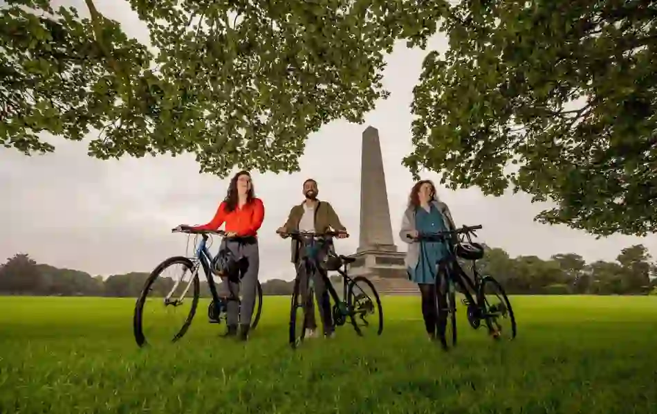 Three people with bicycles in Phoenix Park, Dublin, with the Wellington Monument in view.