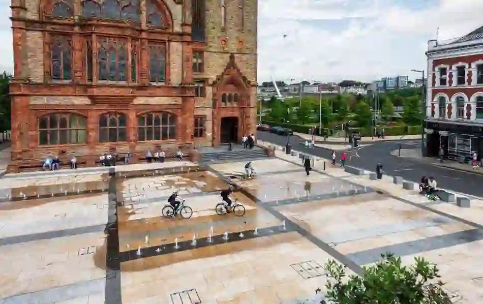 Cyclists riding across Guildhall Square in Derry~Londonderry with fountains and red-brick Guildhall.