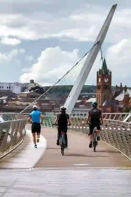 Cyclists crossing the Peace Bridge with Derry~Londonderry’s Guildhall clock tower visible.
