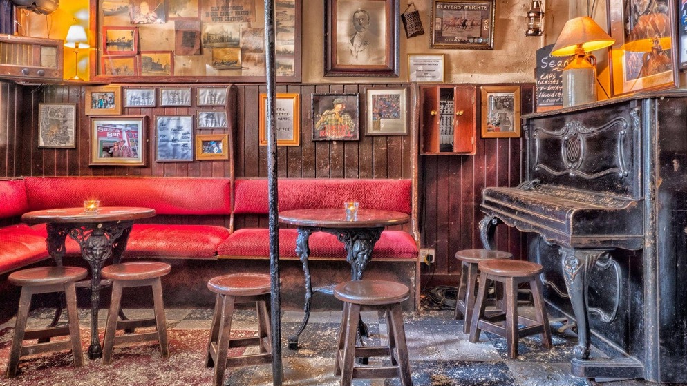 Red-upholstered pub seating with small round tables, stools, framed photos and an old piano.
