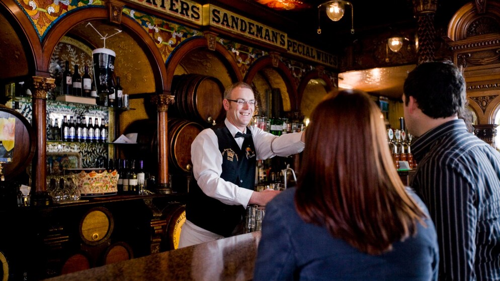 Smiling bartender serving drinks in a traditional Irish pub with ornate décor.