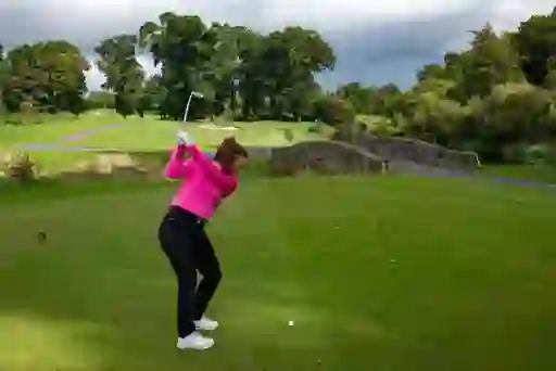 A woman swings her club on a green fairway at The K Club in County Kildare, with a stone bridge and trees behind her.