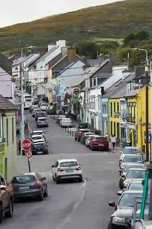 Busy main street in Dingle, lined with colourful pubs, shops, and parked cars against a hillside backdrop.