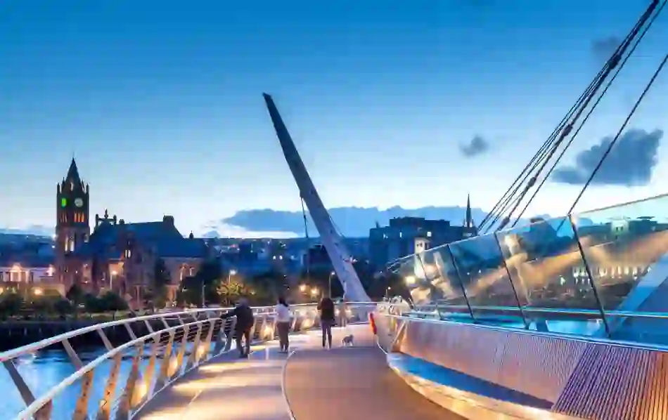 Evening view of the Peace Bridge in Derry~Londonderry, with Guildhall and city skyline lit against the sky.