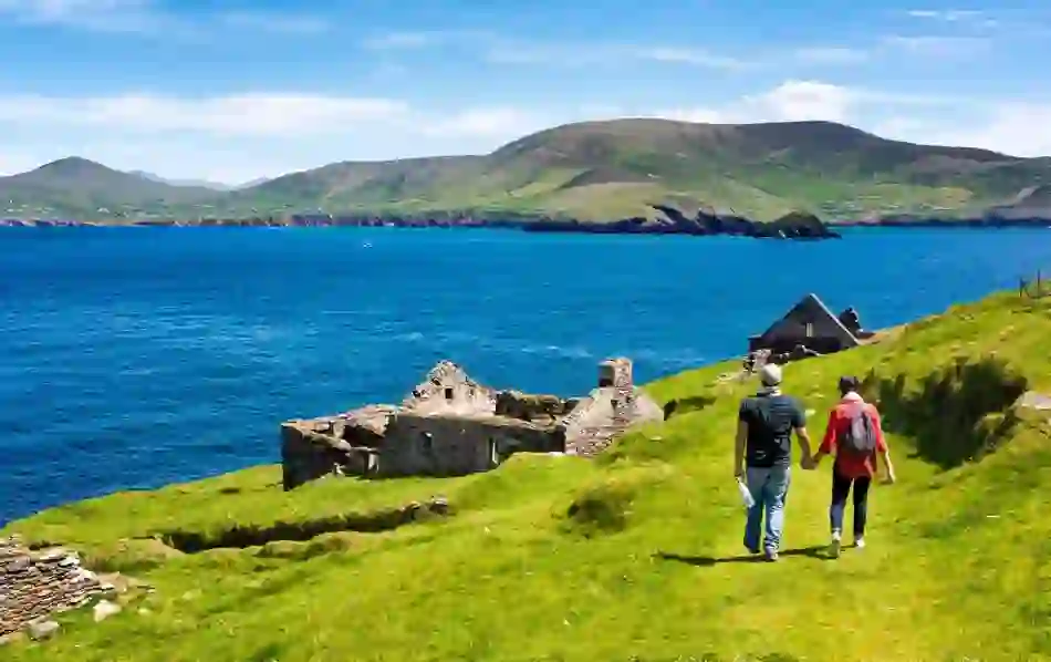 Couple walking hand in hand past stone ruins towards the blue waters off Great Blasket Island, County Kerry.
