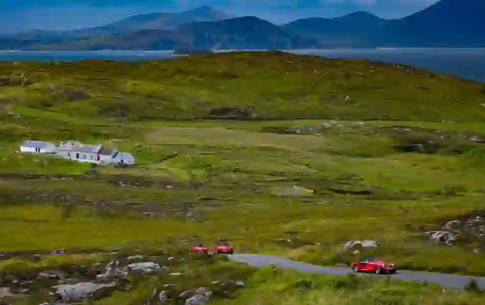 Red classic cars driving along a winding rural road past white cottages with mountains and sea views on the Wild Atlantic Way.