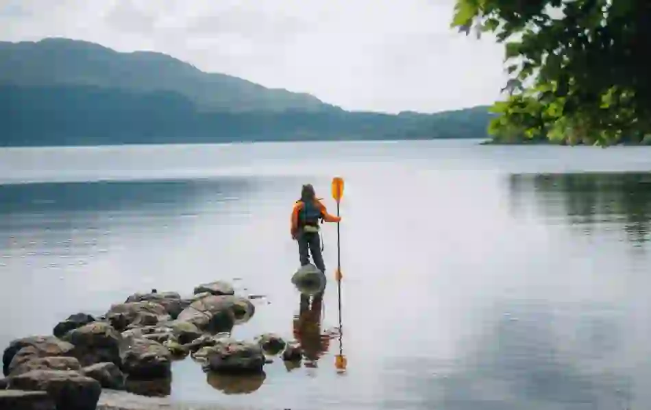 Person in orange jacket holding a paddle while standing on lakeshore rocks in Sligo with mountains in the distance.