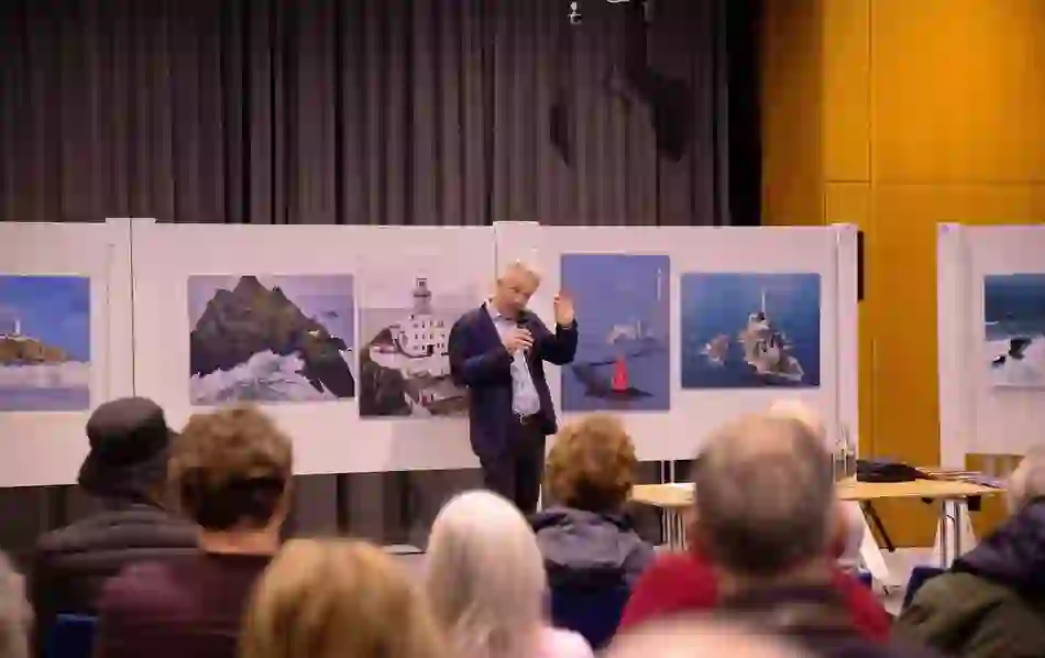 A speaker gestures during a literary talk at the Dublin Book Festival, with lighthouse photos on display behind him.
