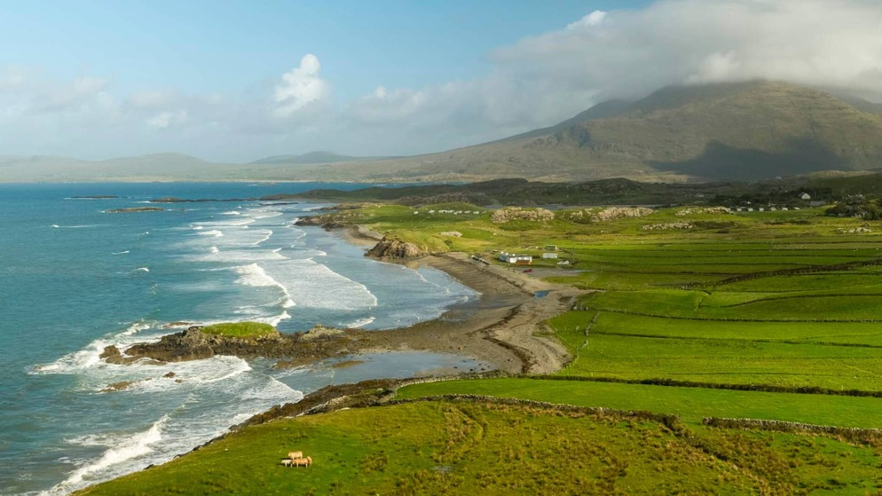 Waves roll in beside green coastal fields beneath misty mountains in the Gaeltacht region of County Mayo.