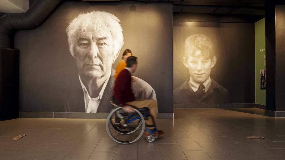 Portraits of Seamus Heaney and his younger self at HomePlace; visitors pass by, immersed in his story.