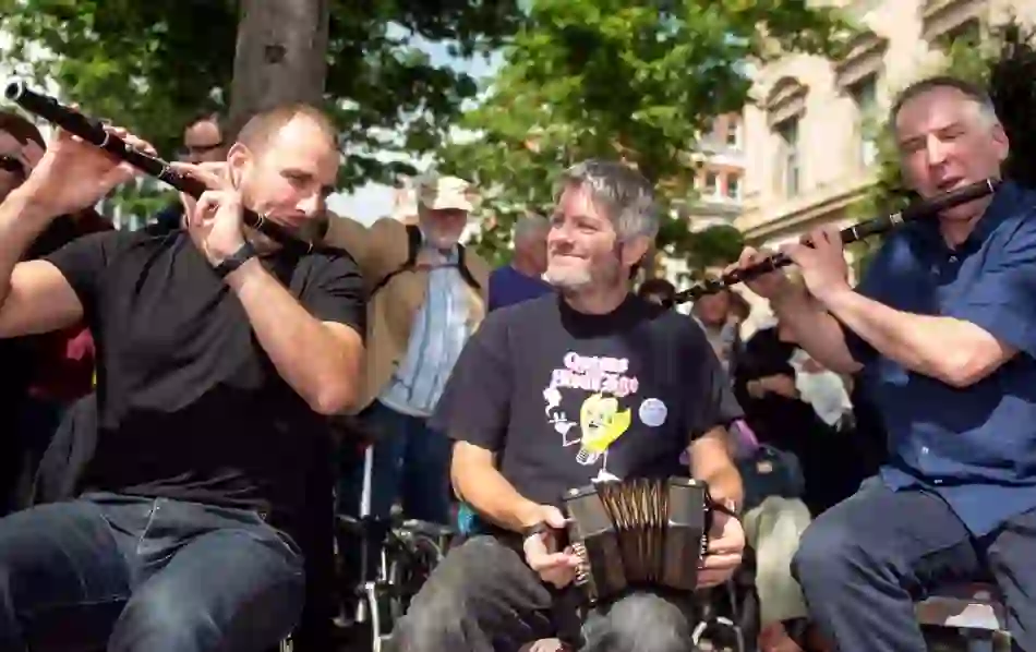 Three musicians play flute and concertina at Fleadh Cheoil, Derry~Londonderry, with a crowd behind them.
