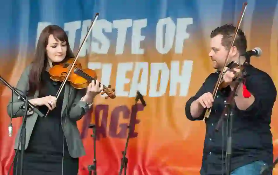 Two fiddlers perform on stage at Fleadh Cheoil, Derry~Londonderry, beneath a backdrop reading 'Taste of Fleadh.'