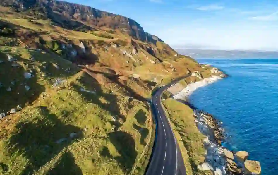 Coastal road on Causeway Coastal Route, Antrim, curving beside rocky cliffs and blue sea waters.