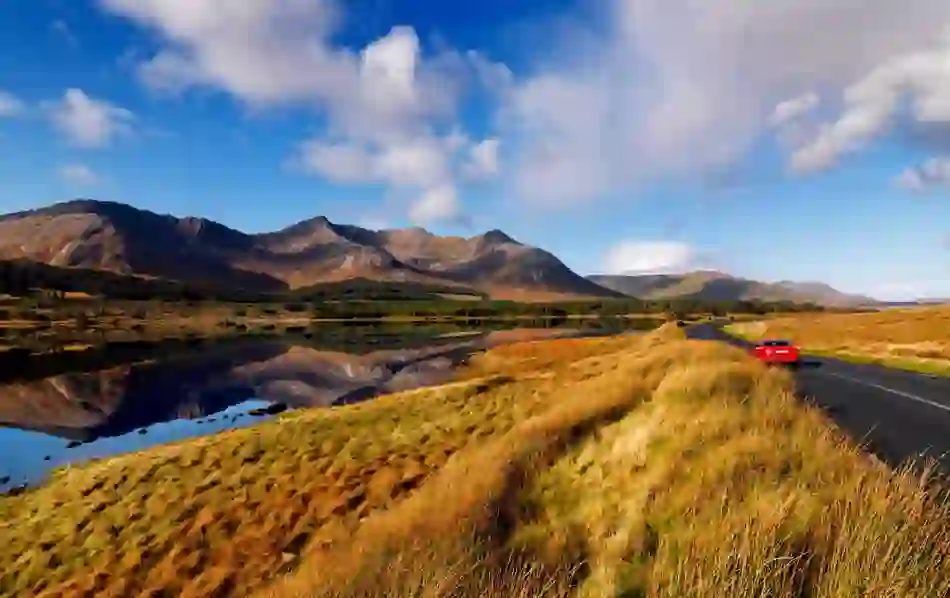 Red car driving past Twelve Bens mountains in Connemara, Galway, reflected in a calm lakeside view.