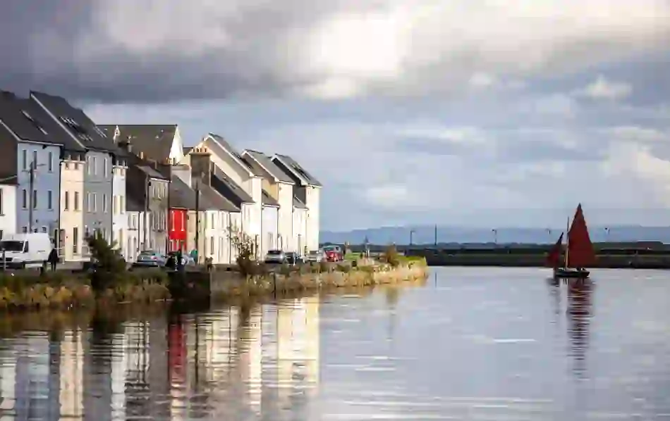 Traditional Galway Hooker boat with red sails passing colourful houses along the Claddagh quay in Galway City.