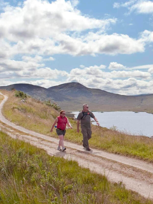 Two walkers follow a winding trail along Lough Veagh in County Donegal, with rugged hills under a cloud-filled summer sky.