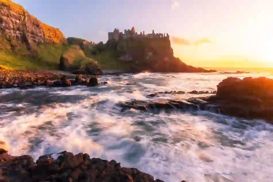Waves surge against black rocks beneath the ruins of a cliffside castle at sunset.