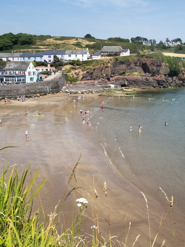 Crowded beach in Dunmore East, County Waterford, with people swimming and colourful houses lining the shore.