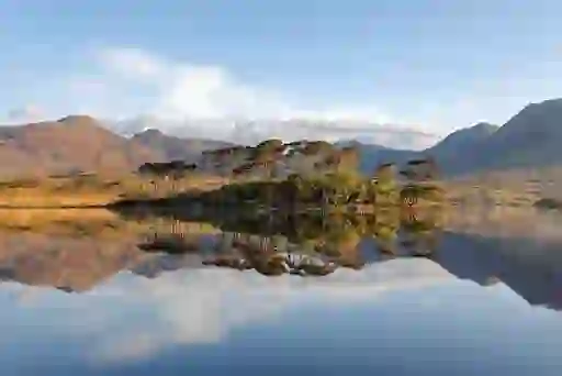 Small pine-covered island reflected in calm waters of Derryclare Lough, with Connemara mountains under soft evening light.