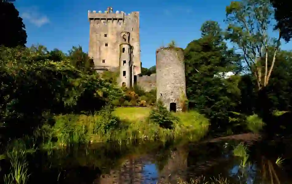 View of medieval Blarney Castle and tower reflected in a pond, surrounded by trees in County Cork.