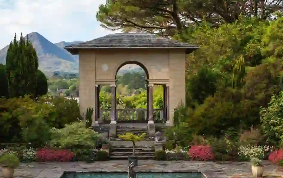 A formal garden on Garnish Island, Cork, featuring an elegant stone archway, manicured flowerbeds, and mountains in the background.