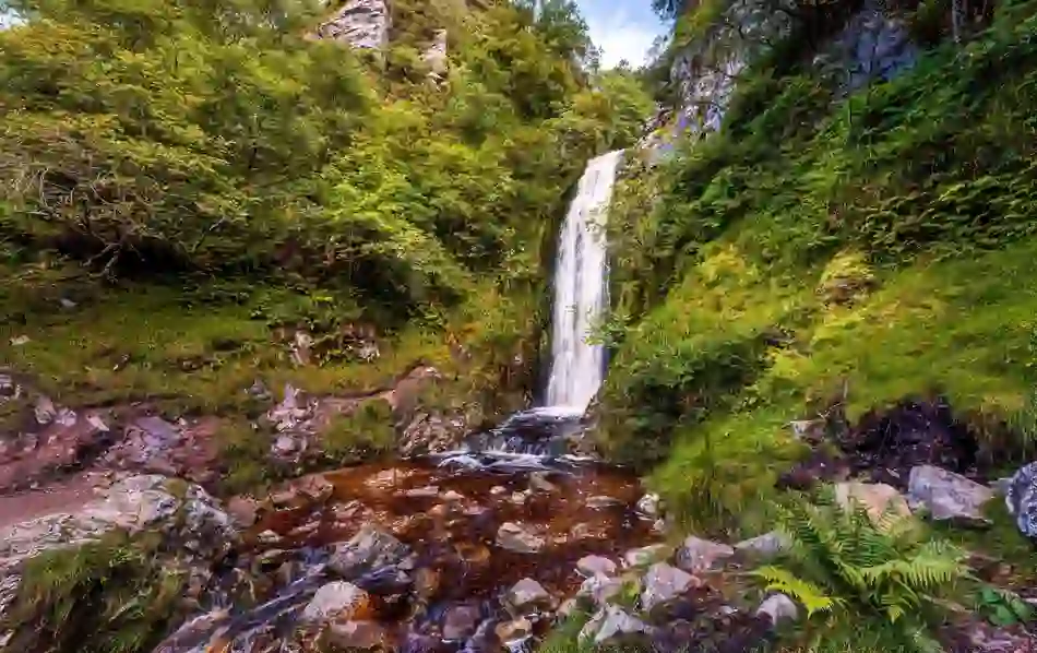 A picturesque waterfall cascades into a rocky pool surrounded by lush green foliage at Glenevin, Donegal.