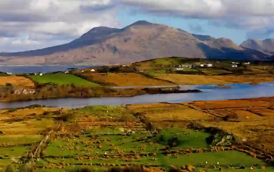 A patchwork of green and golden fields in Uggool Valley, Mayo, with mountains rising behind and a lake reflecting the landscape.