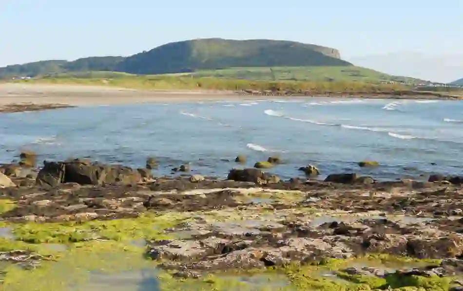 A view of the sea and rocky shore with Queen Maeve’s Cairn, Sligo, visible atop a flat-topped hill in the distance.
