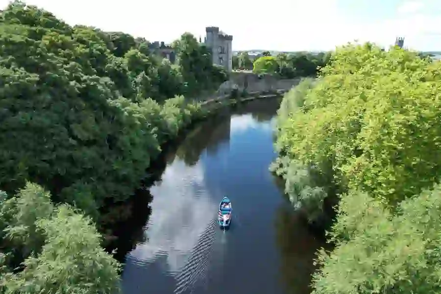 Small blue boat with passengers gliding along the River Nore, flanked by lush greenery and Kilkenny Castle.