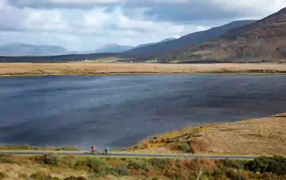 Two cyclists ride a trail beside a vast lake, framed by open bogland and distant mountains.