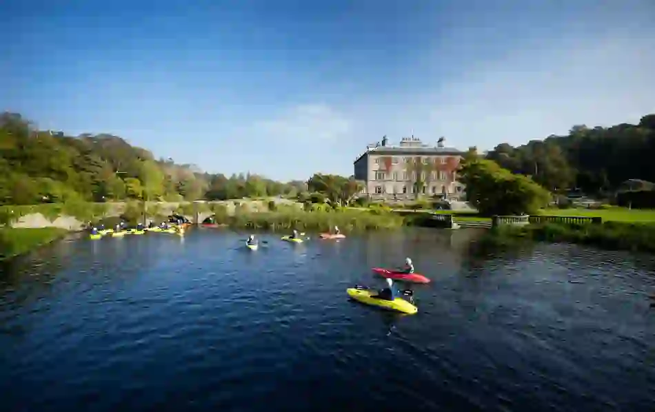 A group of kayakers paddle on calm river waters near a stately home surrounded by green parkland.