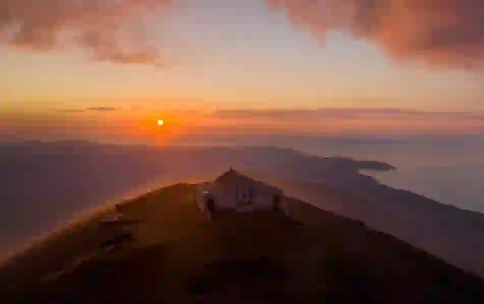 A small white chapel sits atop a mountain ridge at sunset, with golden light spilling across land and sea.