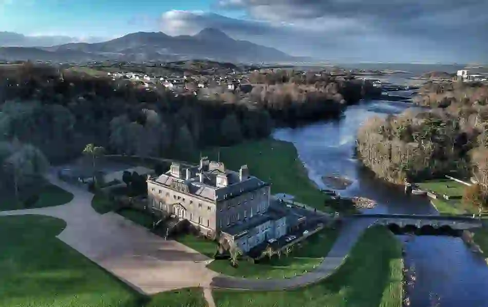 Aerial view of Westport House beside a river, with forest, village rooftops, and Croagh Patrick in the background.