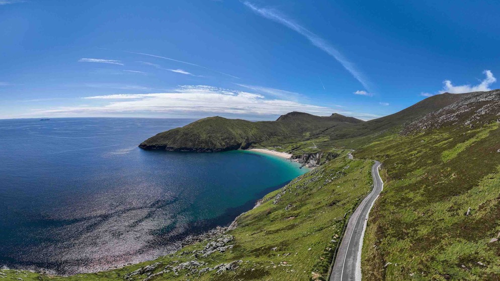 Coastal road curving along cliffs above the Atlantic Ocean on Ireland’s Wild Atlantic Way, under blue sky with wispy clouds.