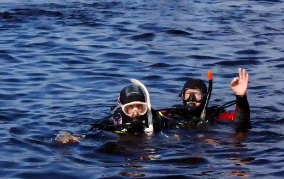 Two scuba divers in wetsuits and masks float in dark water at Dromineer, County Tipperary, one signalling OK with hand.