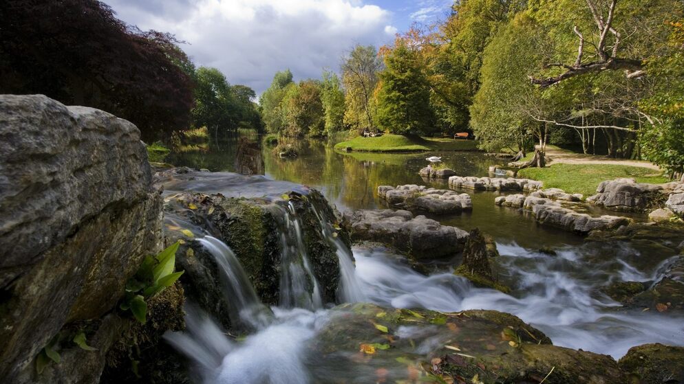 Small waterfall flows into a calm pond surrounded by trees and gardens in Ireland.