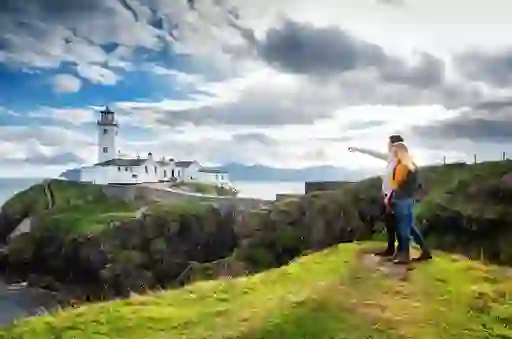 Two people stand on a grassy cliff pointing toward Fanad Lighthouse on Donegal’s dramatic Atlantic coastline.
