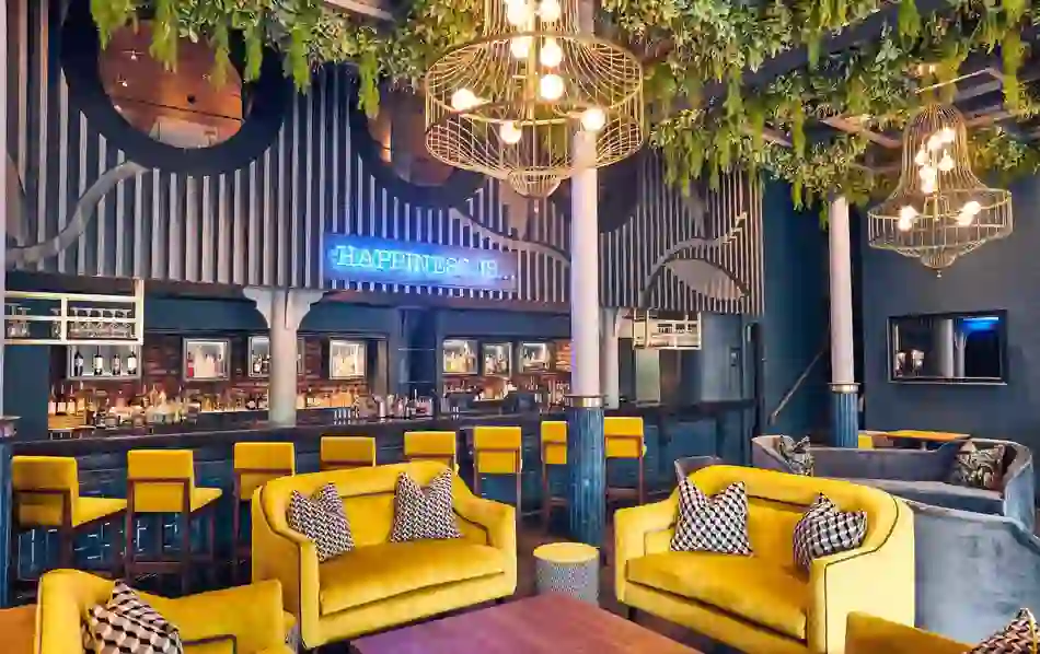 Lush cocktail bar with yellow armchairs, hanging plants and a neon ‘Happiness Is…’ sign in Malmaison, Belfast.