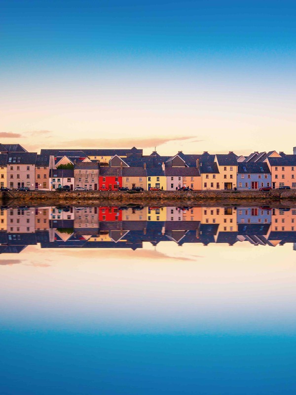 Row of pastel-coloured houses reflected in the water at Galway’s Long Walk during sunset.