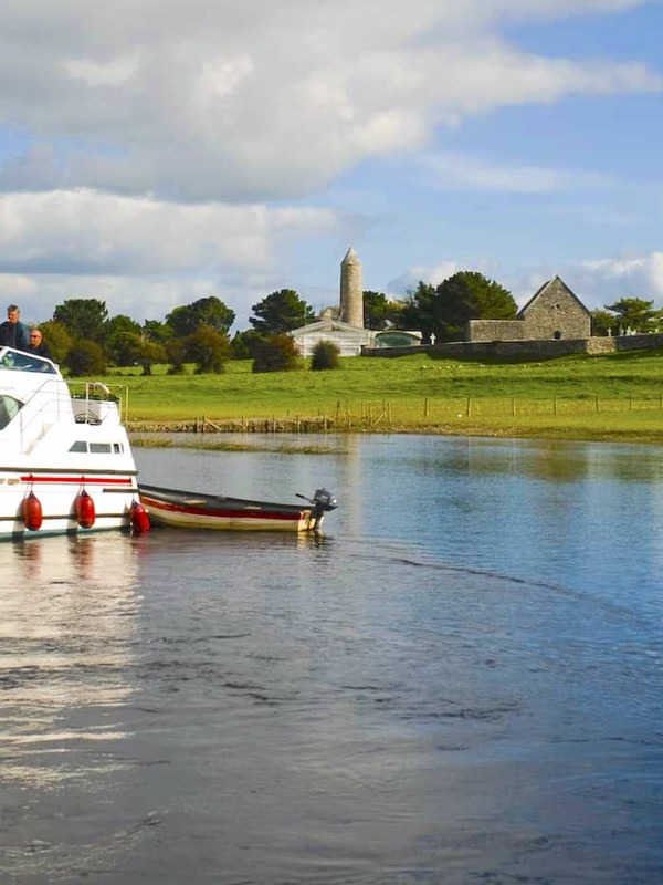 White motor cruiser on the River Shannon with Clonmacnoise monastery and round tower in the background.