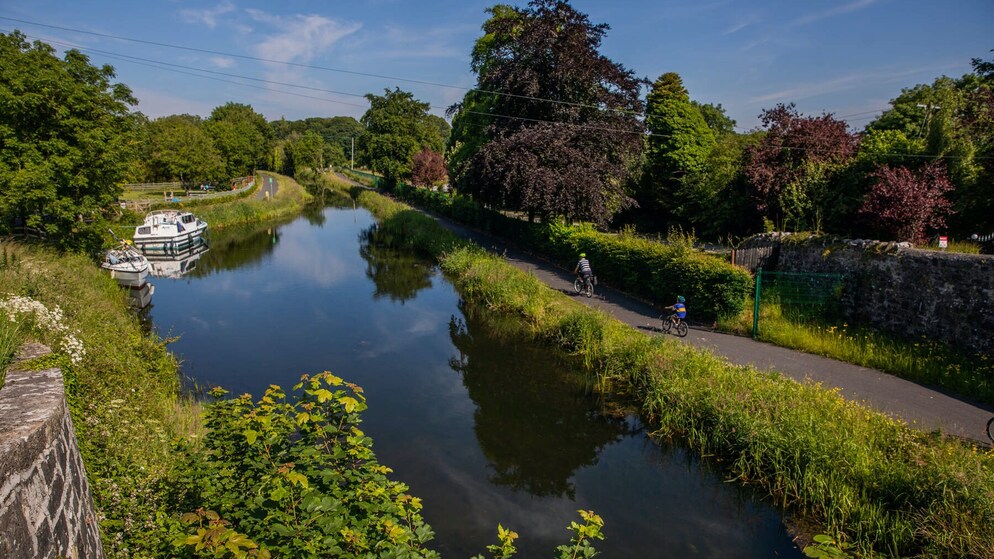 Two cyclists ride along a towpath beside the Royal Canal, with trees, hedgerows and moored boats reflected in the water.