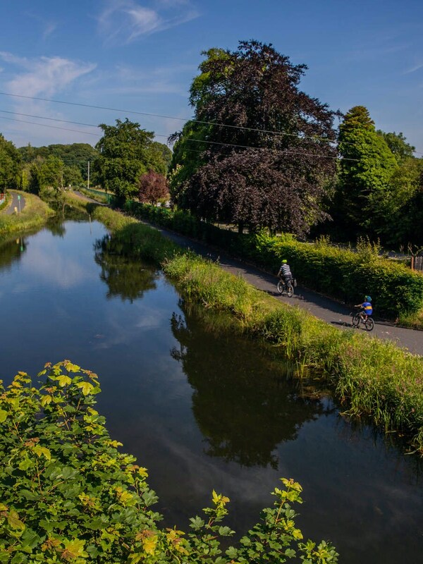 Two cyclists ride along a towpath beside the Royal Canal, with trees, hedgerows and moored boats reflected in the water.