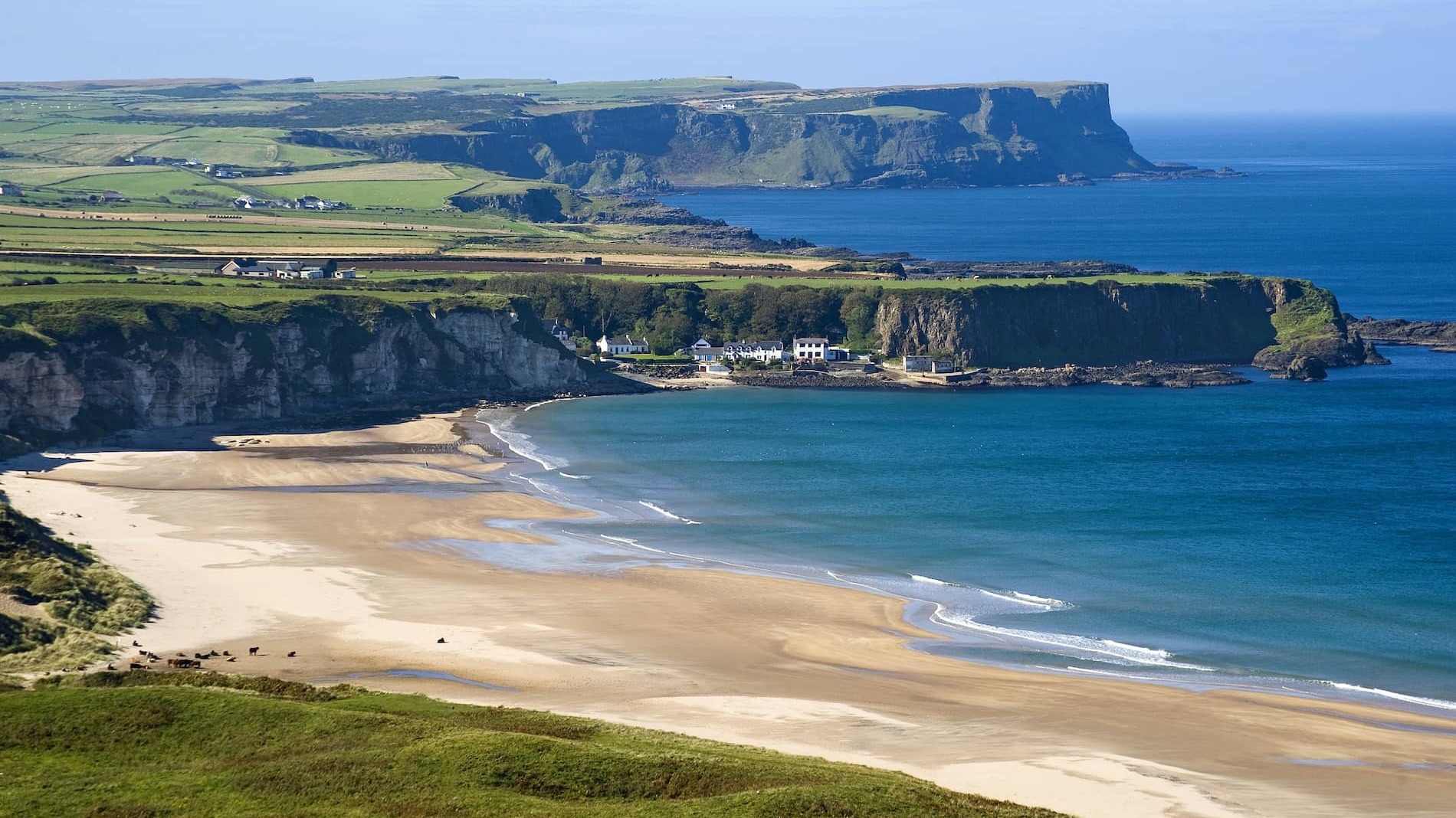 Panoramic view of Whitepark Bay with turquoise sea and white cottages nestled under cliffs, County Antrim.