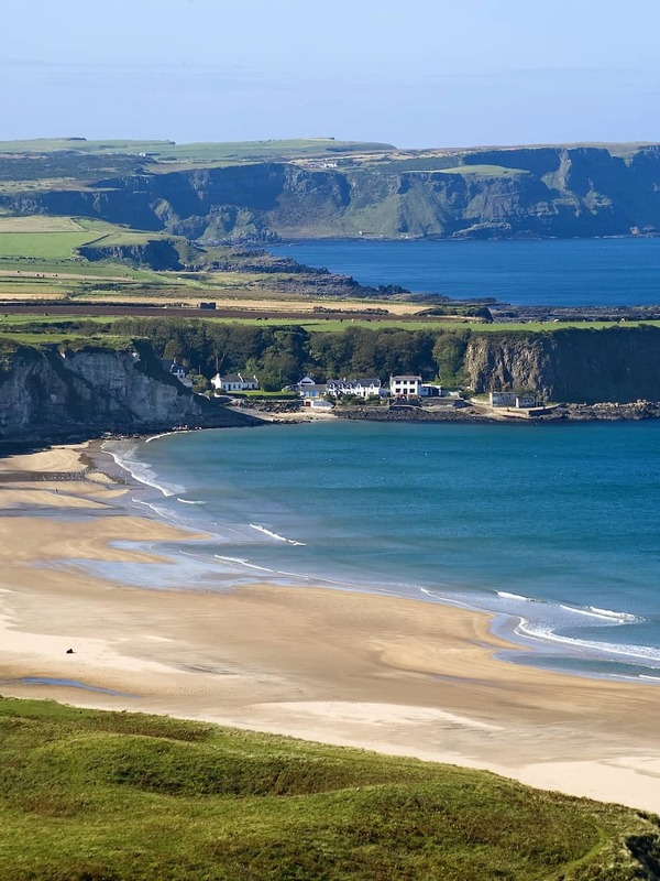 Panoramic view of Whitepark Bay with turquoise sea and white cottages nestled under cliffs, County Antrim.
