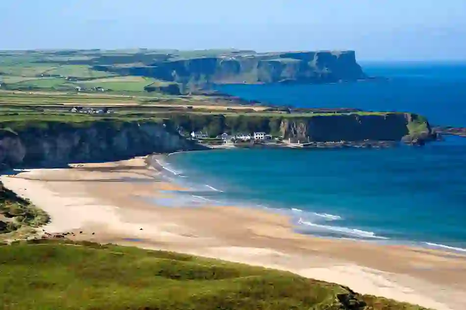 Panoramic view of Whitepark Bay with turquoise sea and white cottages nestled under cliffs, County Antrim.