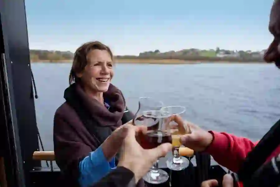 Woman smiling on boat as glasses of wine and juice are raised in a toast on Lough Erne, with shoreline in the distance.