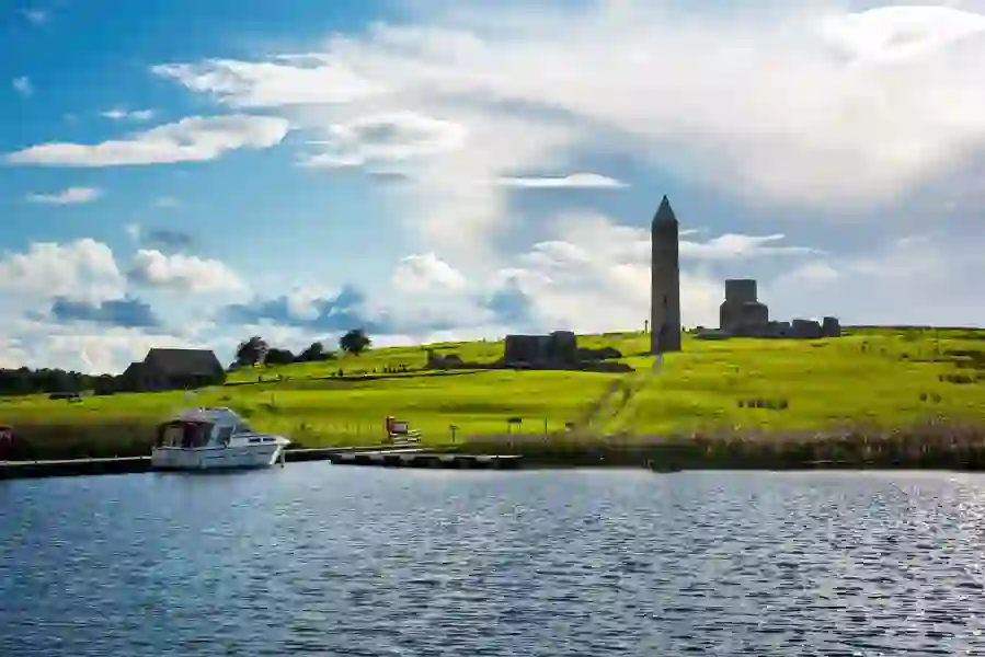 Boat docked at Devenish Island near medieval ruins and tall stone round tower set against rolling green fields and sky.